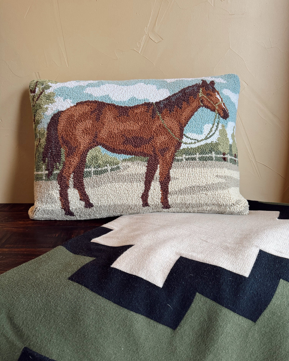 A rectangular needlepoint pillow featuring a brown horse in a field under a blue sky, displayed on a dark wooden surface against a beige wall, with a green and white chevron blanket in the foreground.