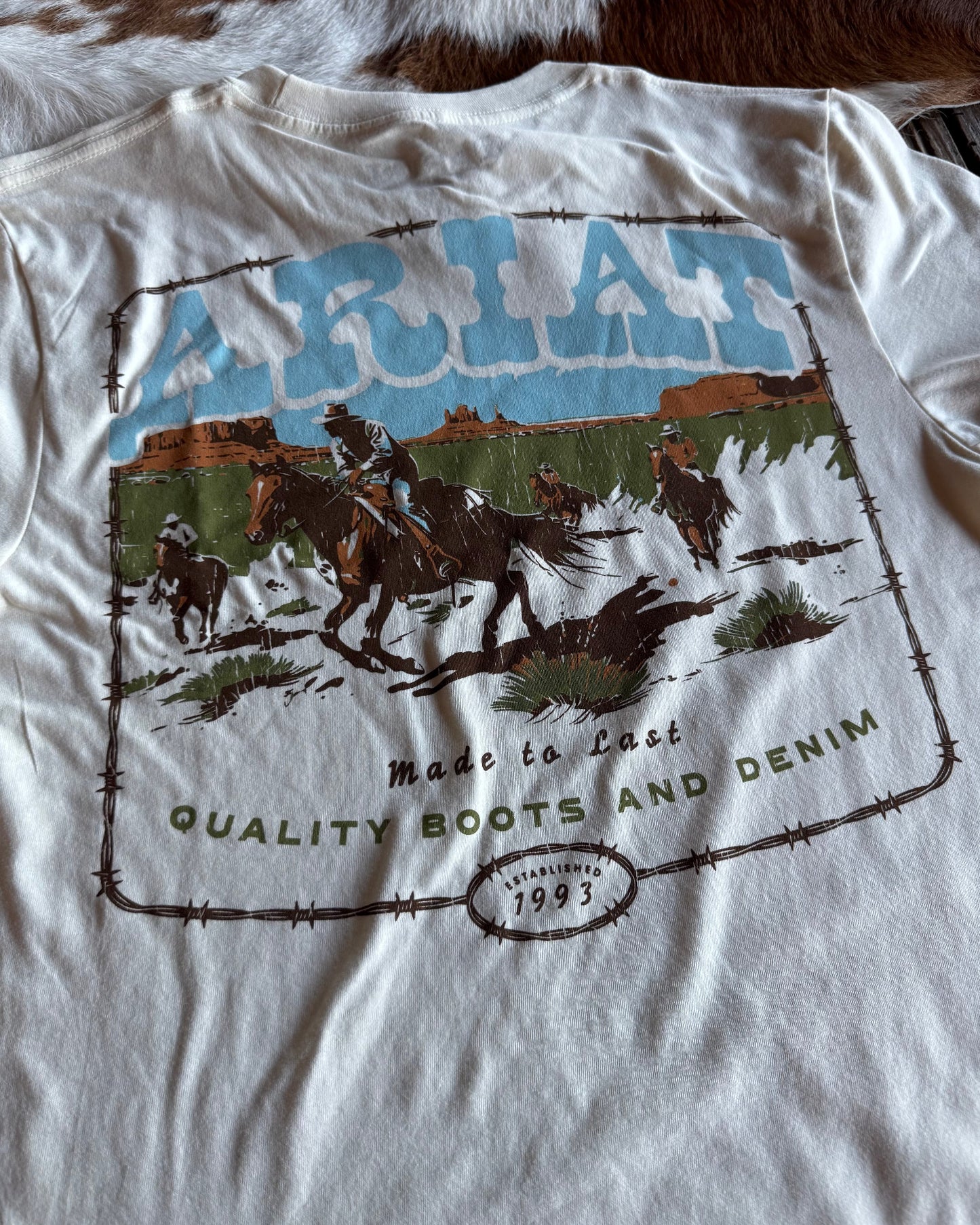 White t-shirt with a graphic design of horses and text on a cowhide rug background