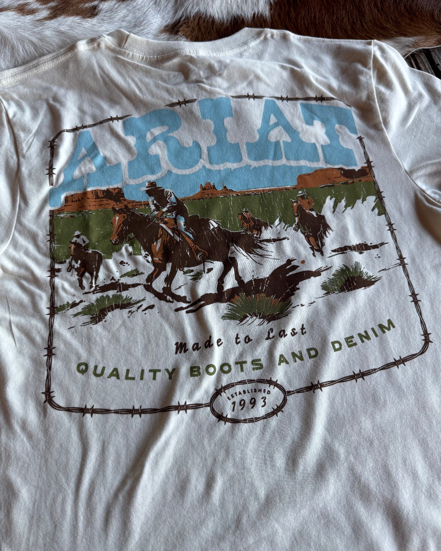 White t-shirt with a graphic design of horses and text on a cowhide rug background