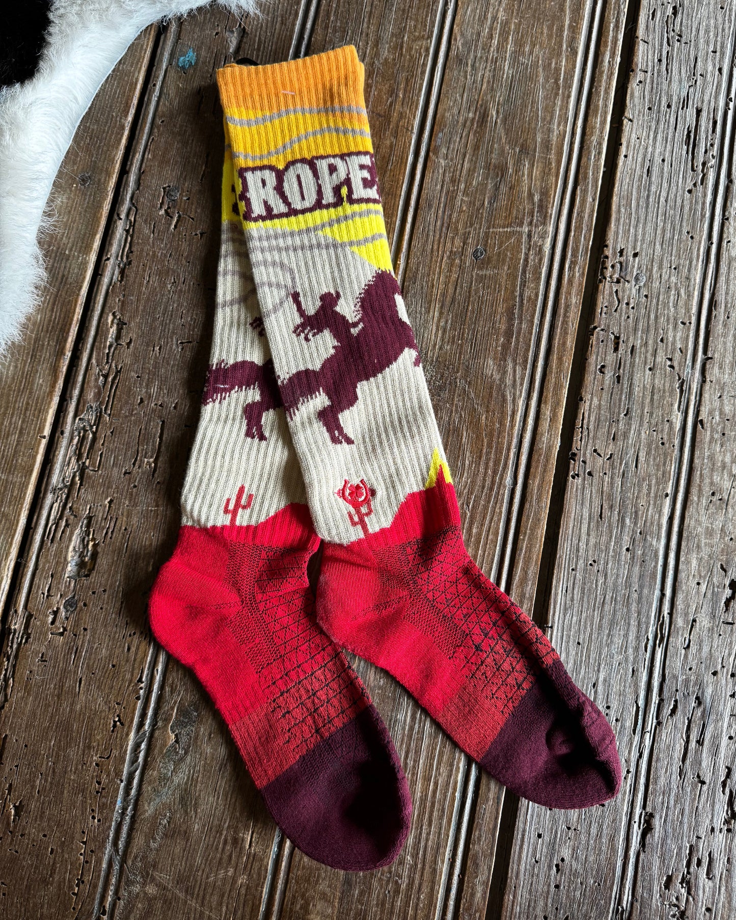 A pair of long knit socks laid flat on a rustic wooden surface. The socks have a cream upper section with a dark burgundy cowboy riding a horse graphic and the word 'ROPE' across the calf, a yellow stripe near the top, and a bright red lower section with darker burgundy toes and heels.