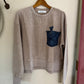 Beige sweatshirt with a blue pocket detail on a hanger against a white wall.