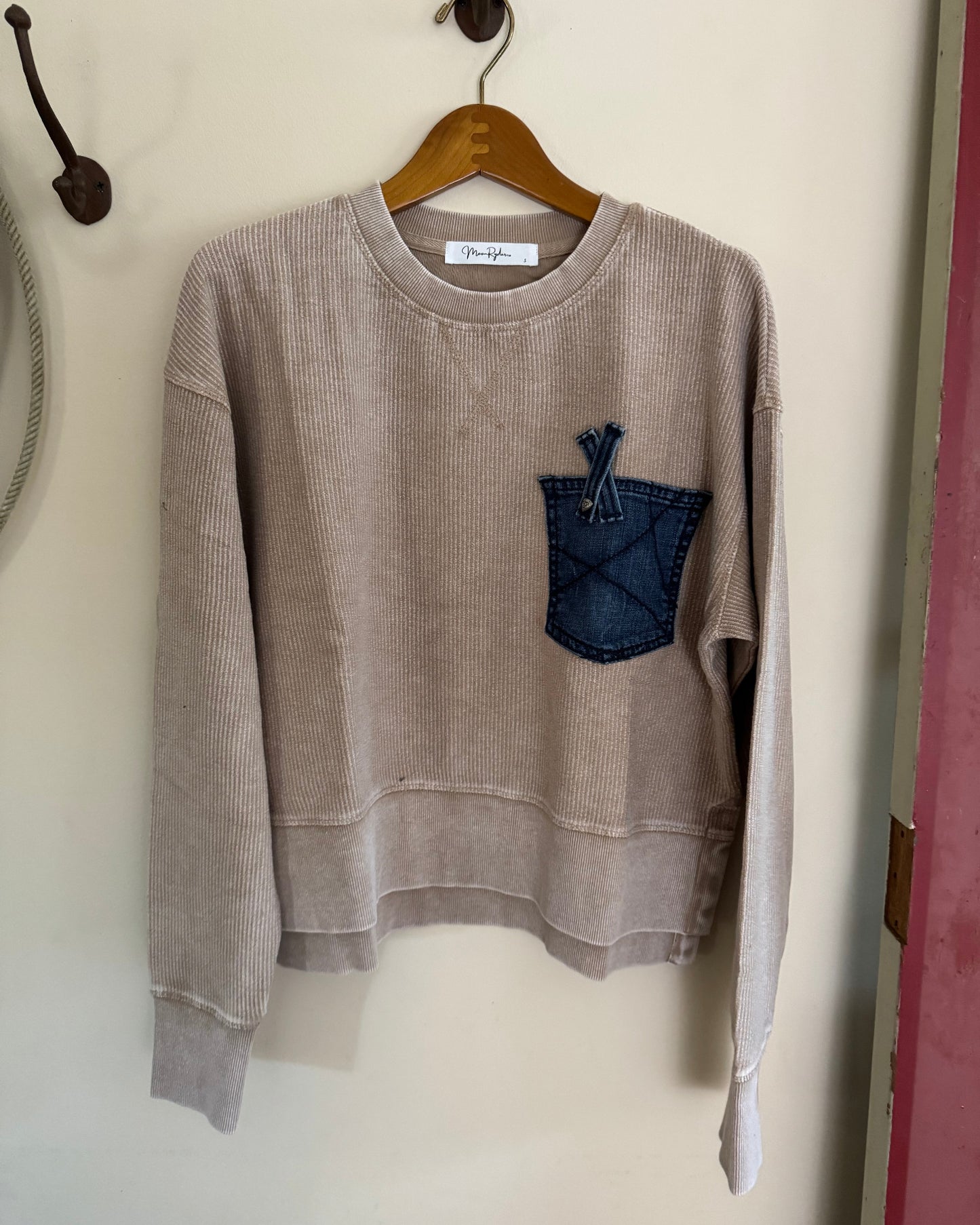 Beige sweatshirt with a blue pocket detail on a hanger against a white wall.