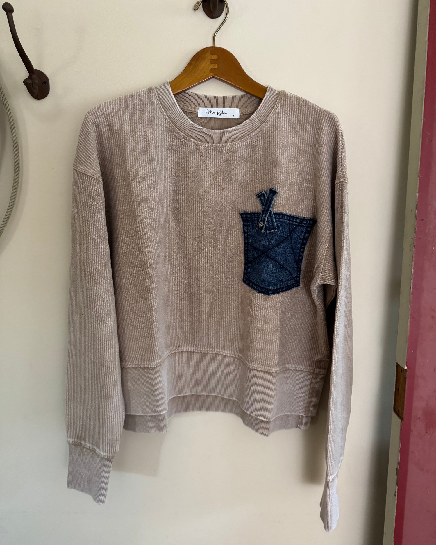 Beige sweatshirt with a blue pocket detail on a hanger against a white wall.