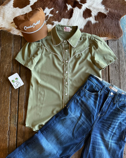 Green shirt, blue jeans, and brown cap on a wooden surface with cowhide rug
