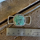 A silver-toned ring with a round green turquoise stone and twisted metal loops, shown on a rustic wooden surface next to a metal ruler for scale.