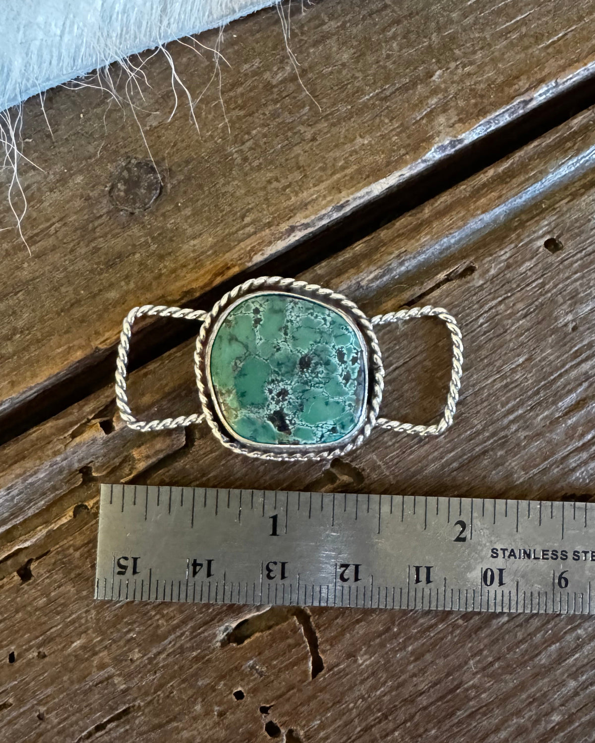 A silver-toned ring with a round green turquoise stone and twisted metal loops, shown on a rustic wooden surface next to a metal ruler for scale.