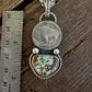 A silver-toned pendant necklace displayed on a rustic wooden surface next to a metal ruler for scale. The pendant features a round silver coin medallion and a heart-shaped turquoise stone with brown matrix.