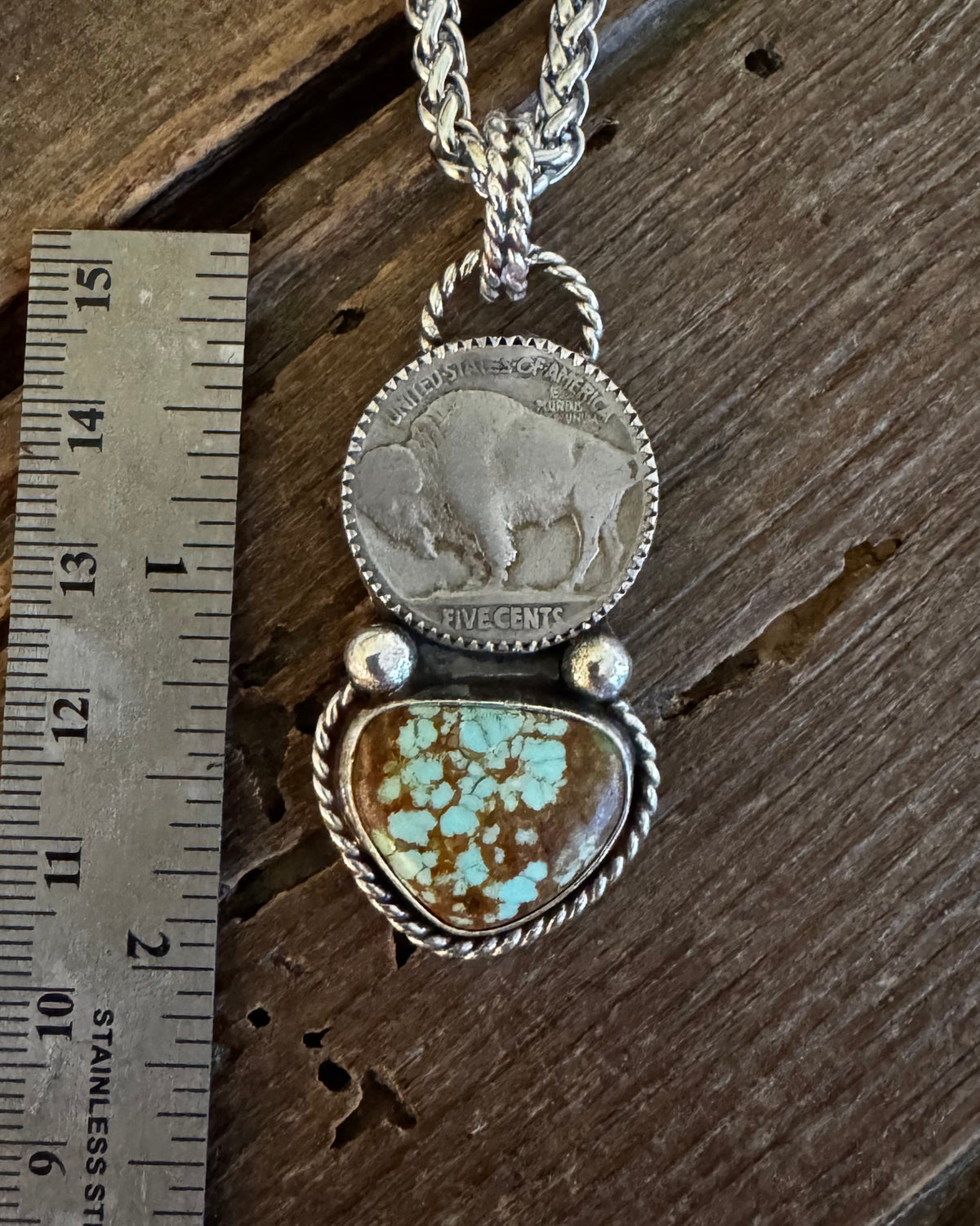 A silver-toned pendant necklace displayed on a rustic wooden surface next to a metal ruler for scale. The pendant features a round silver coin medallion and a heart-shaped turquoise stone with brown matrix.