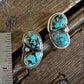 A pair of silver stud earrings with three oval turquoise stones each, displayed on a rustic wooden surface next to a metal ruler for scale.