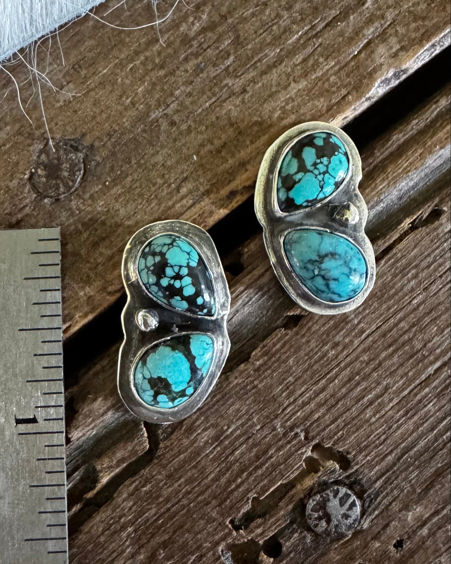 A pair of silver stud earrings with three oval turquoise stones each, displayed on a rustic wooden surface next to a metal ruler for scale.