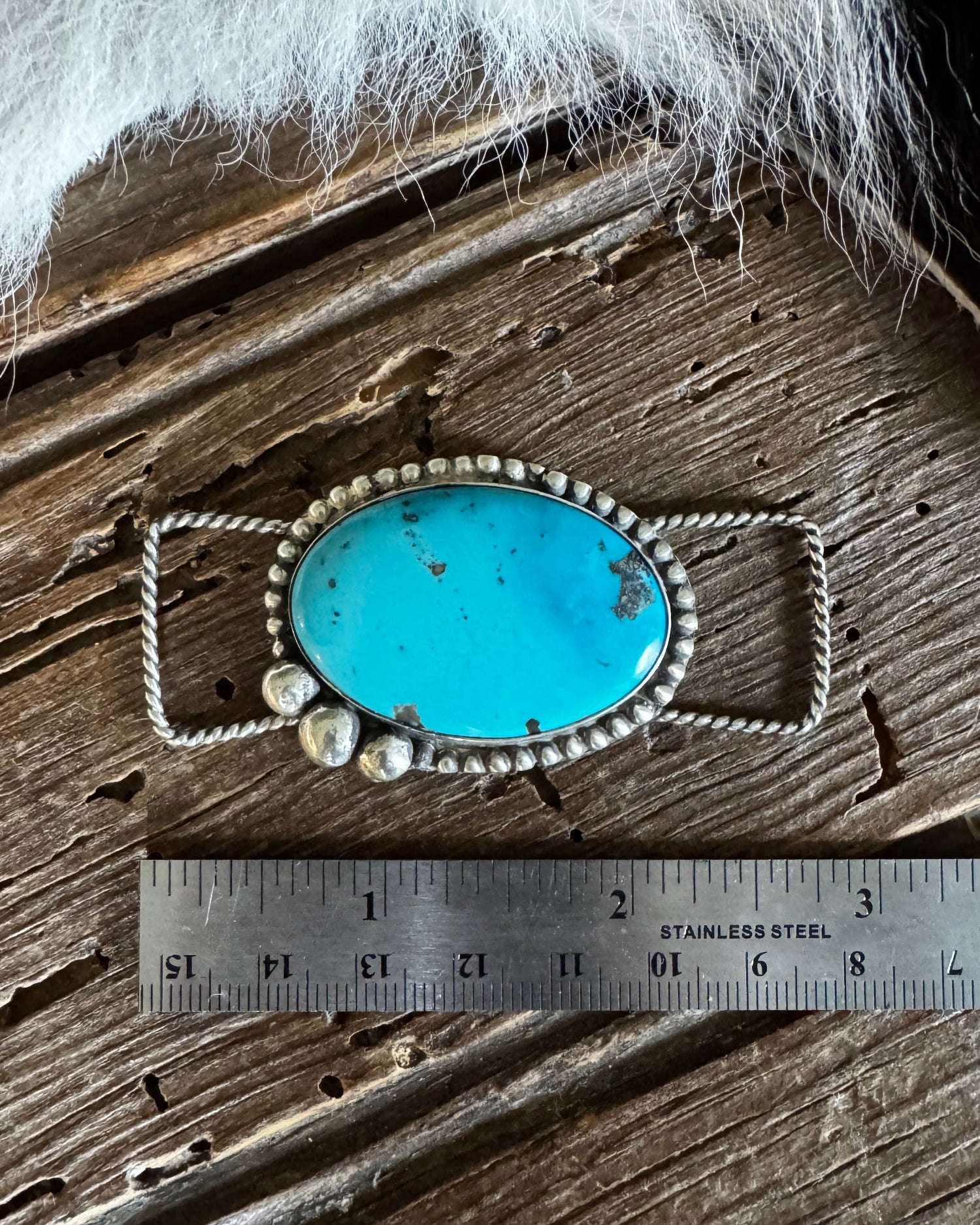 Turquoise pendant with silver frame and pearls on a wooden surface with a ruler for scale.