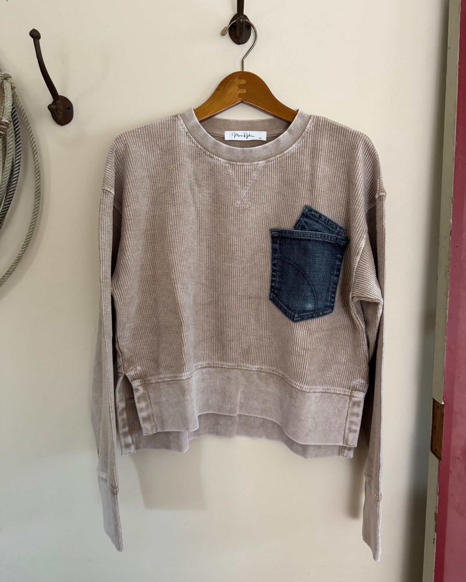 Brown sweatshirt with a denim pocket on a hanger against a white wall.