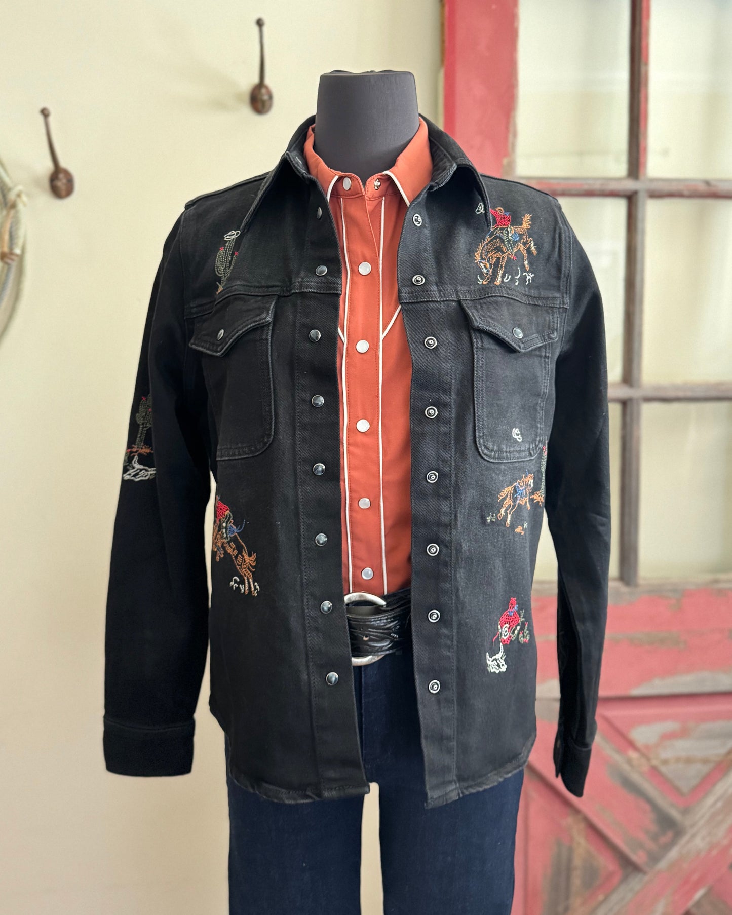 Black denim shirt with embroidered designs on a mannequin against a neutral wall.