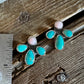 A pair of turquoise and pink stud earrings with silver settings, displayed next to a stainless steel ruler on a rustic wooden surface.