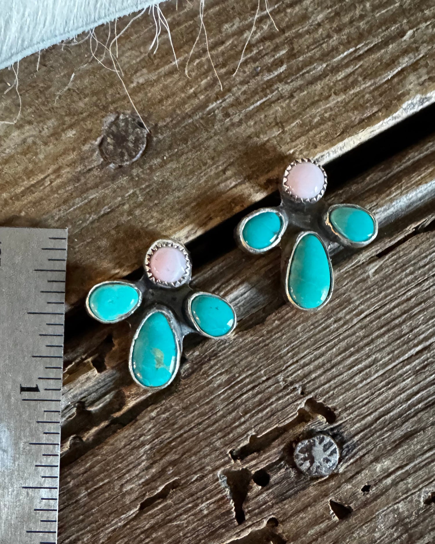 A pair of turquoise and pink stud earrings with silver settings, displayed next to a stainless steel ruler on a rustic wooden surface.