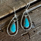 A pair of silver dangle earrings with teardrop-shaped turquoise stones, displayed on a rustic dark wood surface.