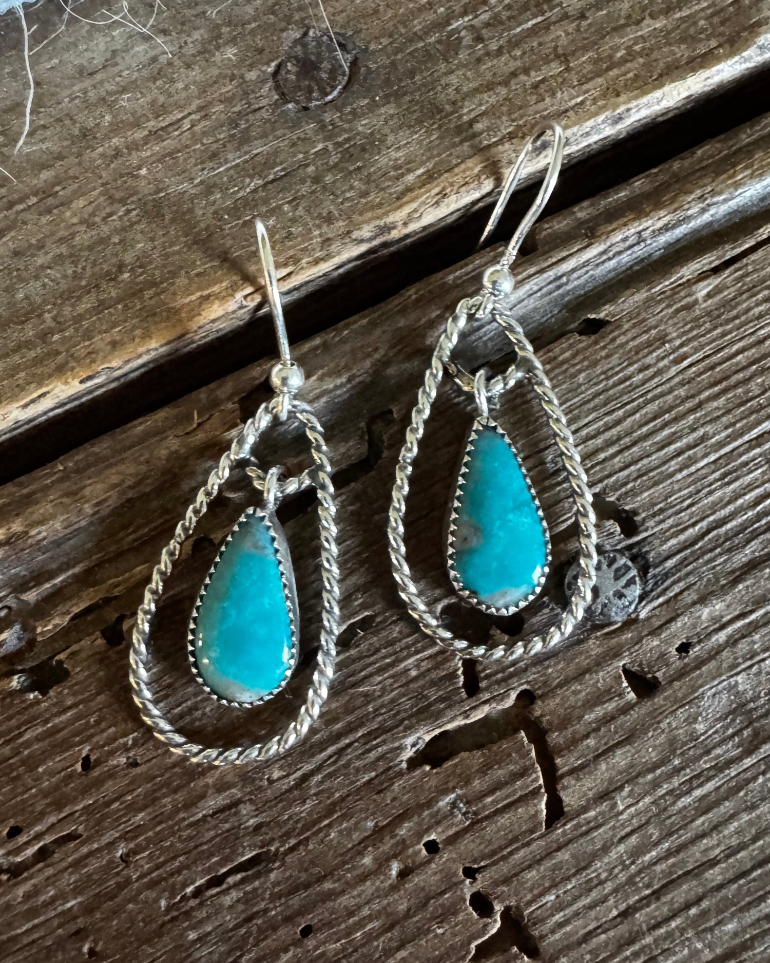 A pair of silver dangle earrings with teardrop-shaped turquoise stones, displayed on a rustic dark wood surface.