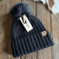Black knit beanie with pom-pom on a wooden surface, featuring a brand tag.
