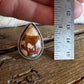A teardrop-shaped cabochon gemstone with a brown and cream marbled pattern, set in a silver-toned bezel with a twisted wire border. The ring is shown on a rustic wooden surface next to a metal ruler held by a hand to indicate its size.