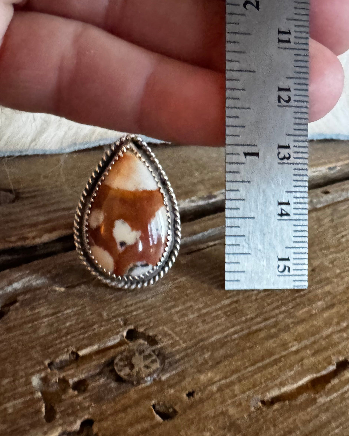 A teardrop-shaped cabochon gemstone with a brown and cream marbled pattern, set in a silver-toned bezel with a twisted wire border. The ring is shown on a rustic wooden surface next to a metal ruler held by a hand to indicate its size.