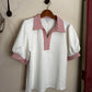 White blouse with red and white striped collar and cuffs on a hanger against a beige wall.