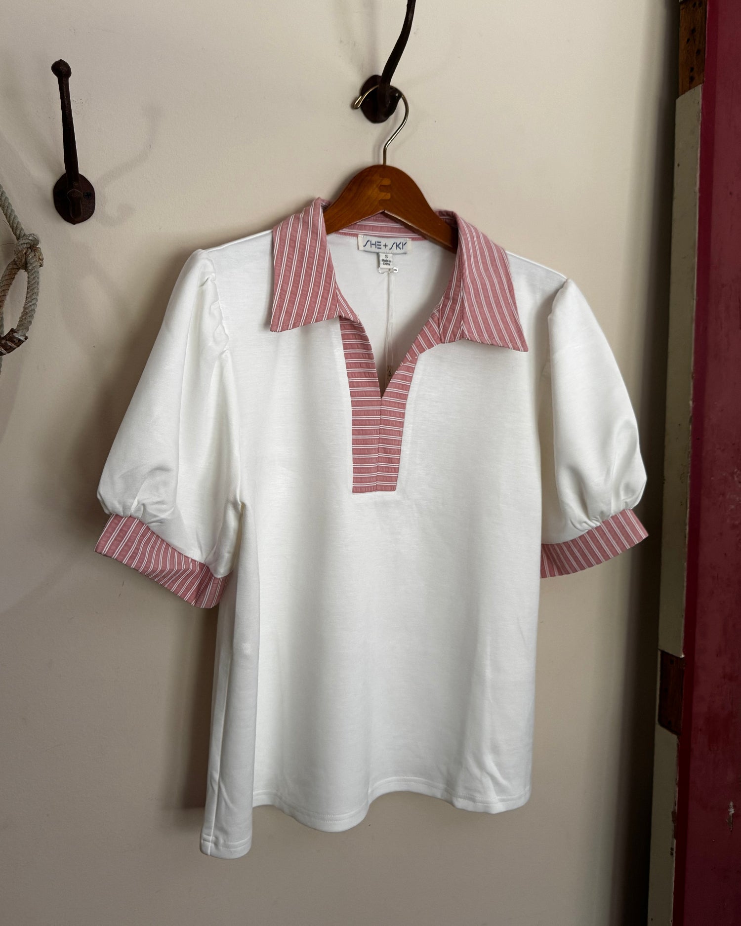White blouse with red and white striped collar and cuffs on a hanger against a beige wall.