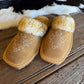 A pair of tan suede slip-on slippers with white faux fur trim and white embroidered western-style floral patterns, displayed on a rustic wooden surface with a black-and-white cowhide rug in the background.
