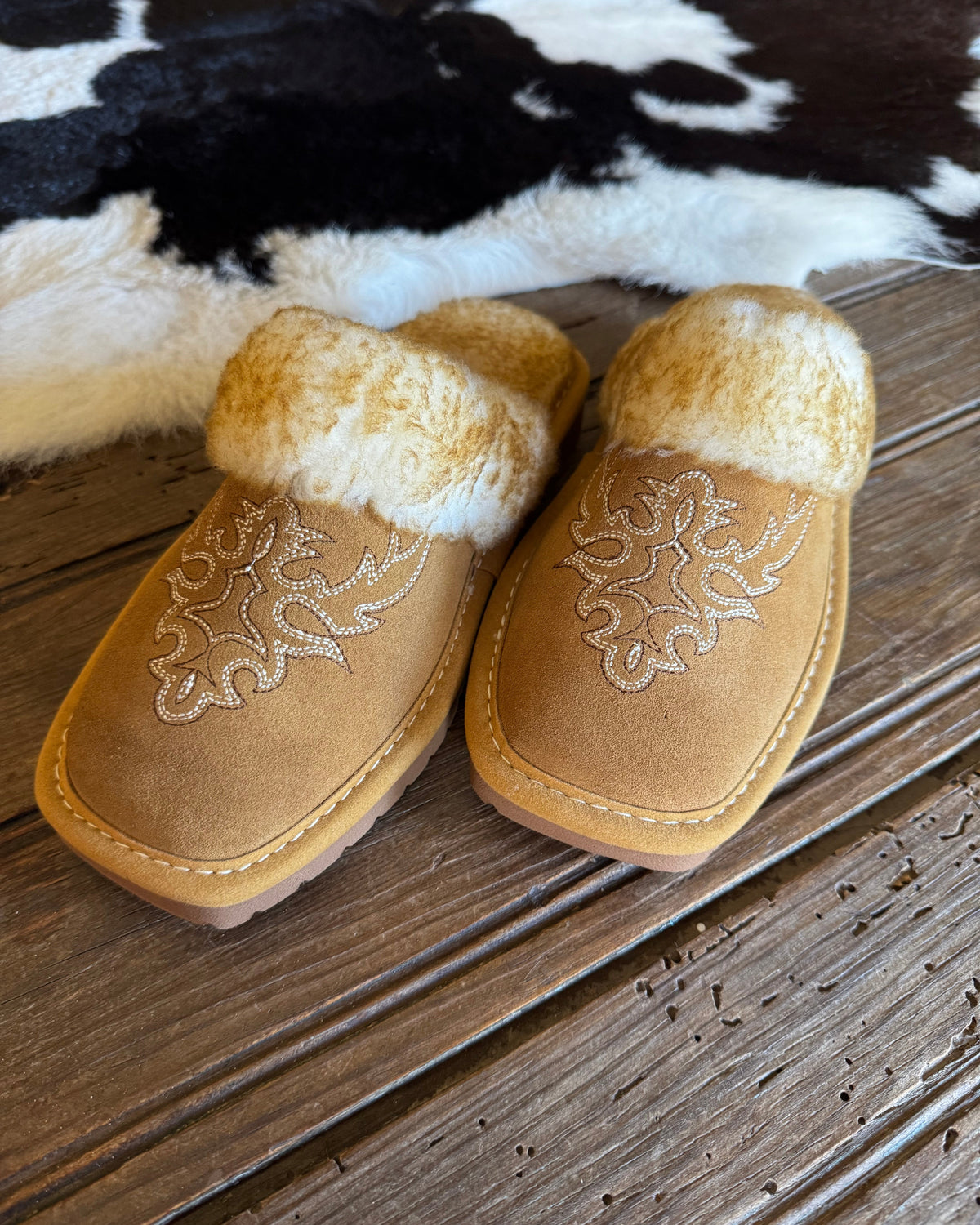 A pair of tan suede slip-on slippers with white faux fur trim and white embroidered western-style floral patterns, displayed on a rustic wooden surface with a black-and-white cowhide rug in the background.