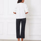 Woman wearing a white blouse and black pants standing against a white wall.