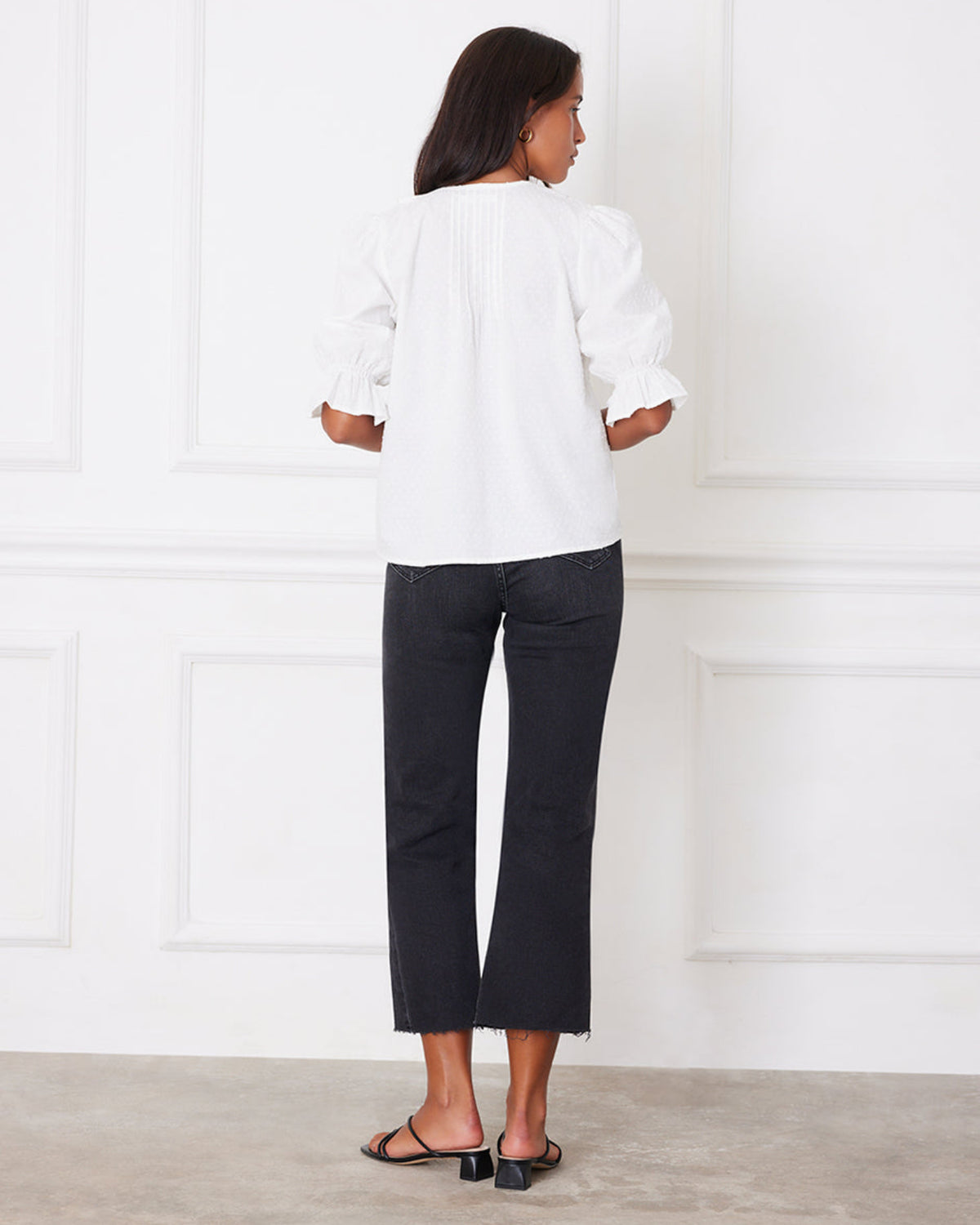 Woman wearing a white blouse and black pants standing against a white wall.