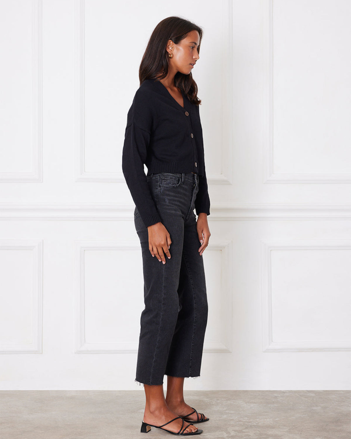 Woman wearing a black cardigan and dark jeans standing against a white paneled wall.