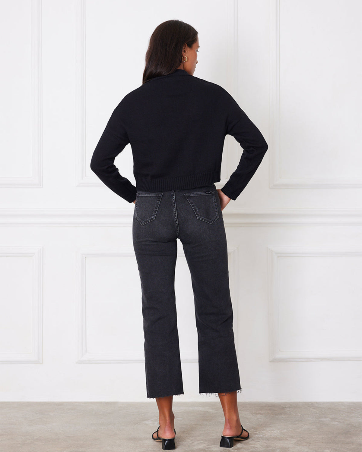 Person wearing a black shirt and dark jeans standing against a white paneled wall.