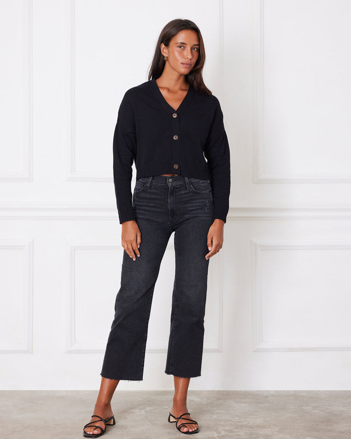 Woman wearing a black cardigan and dark jeans standing against a white paneled wall.