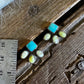 A close-up of several small jewelry pieces laid on a weathered wooden surface, with a metal ruler and white fur visible for scale. The jewelry appears to include turquoise-blue and pale yellow stones in silver-toned settings.