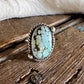 A silver ring with an oval turquoise stone, displayed on a rustic wooden surface with a white fur trim in the background.