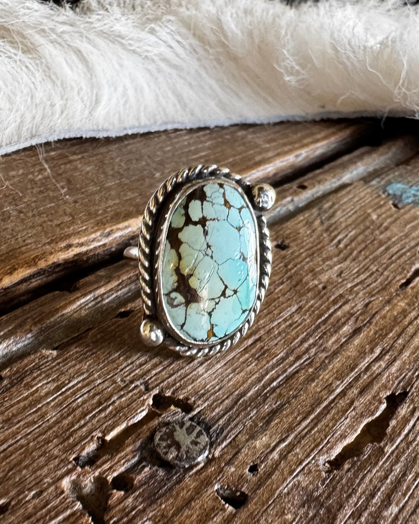 A silver ring with an oval turquoise stone, displayed on a rustic wooden surface with a white fur trim in the background.