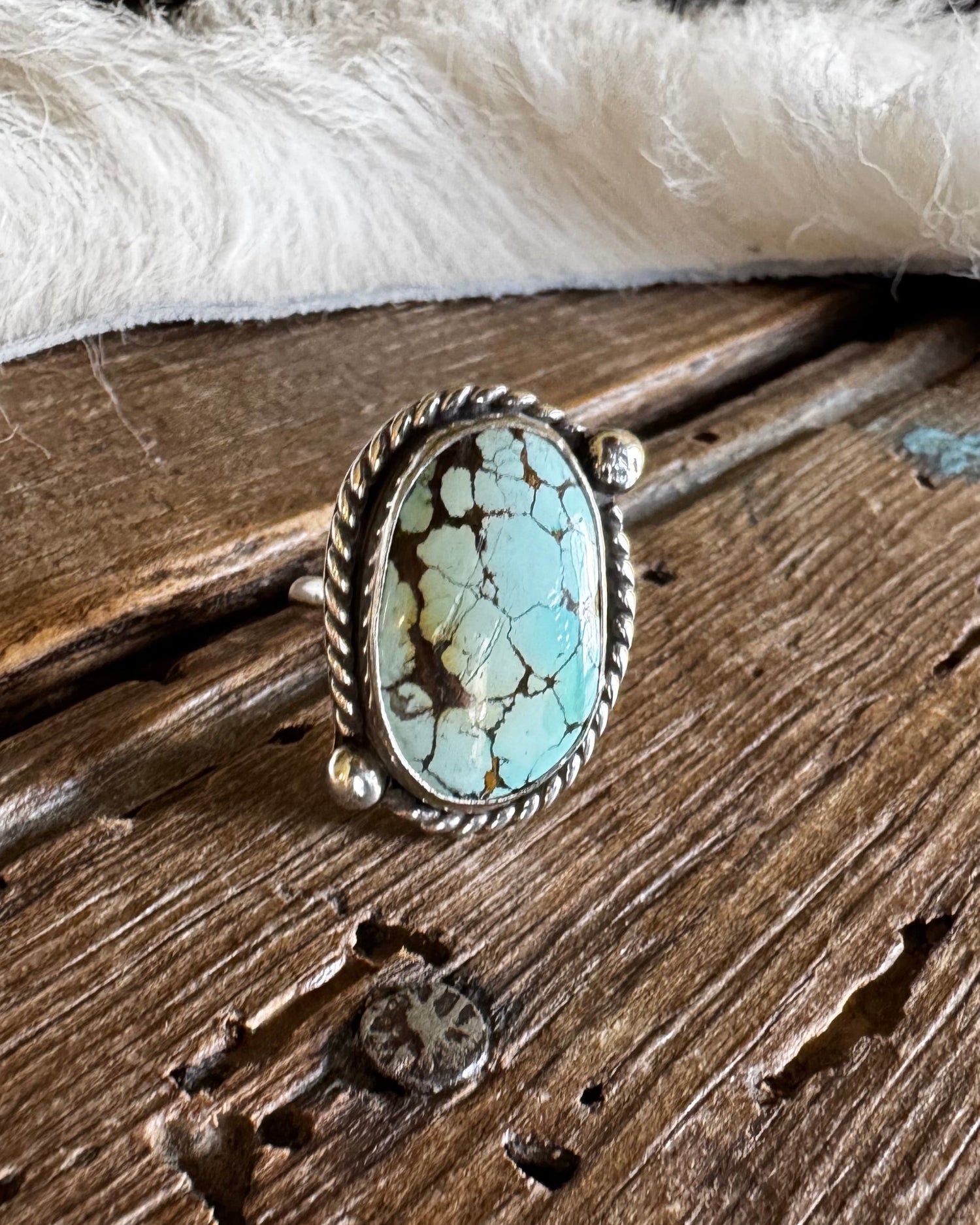 A silver ring with an oval turquoise stone, displayed on a rustic wooden surface with a white fur trim in the background.