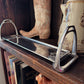 Old cowboy boot, metal stirrup, and ceramic pitcher on a wooden shelf.