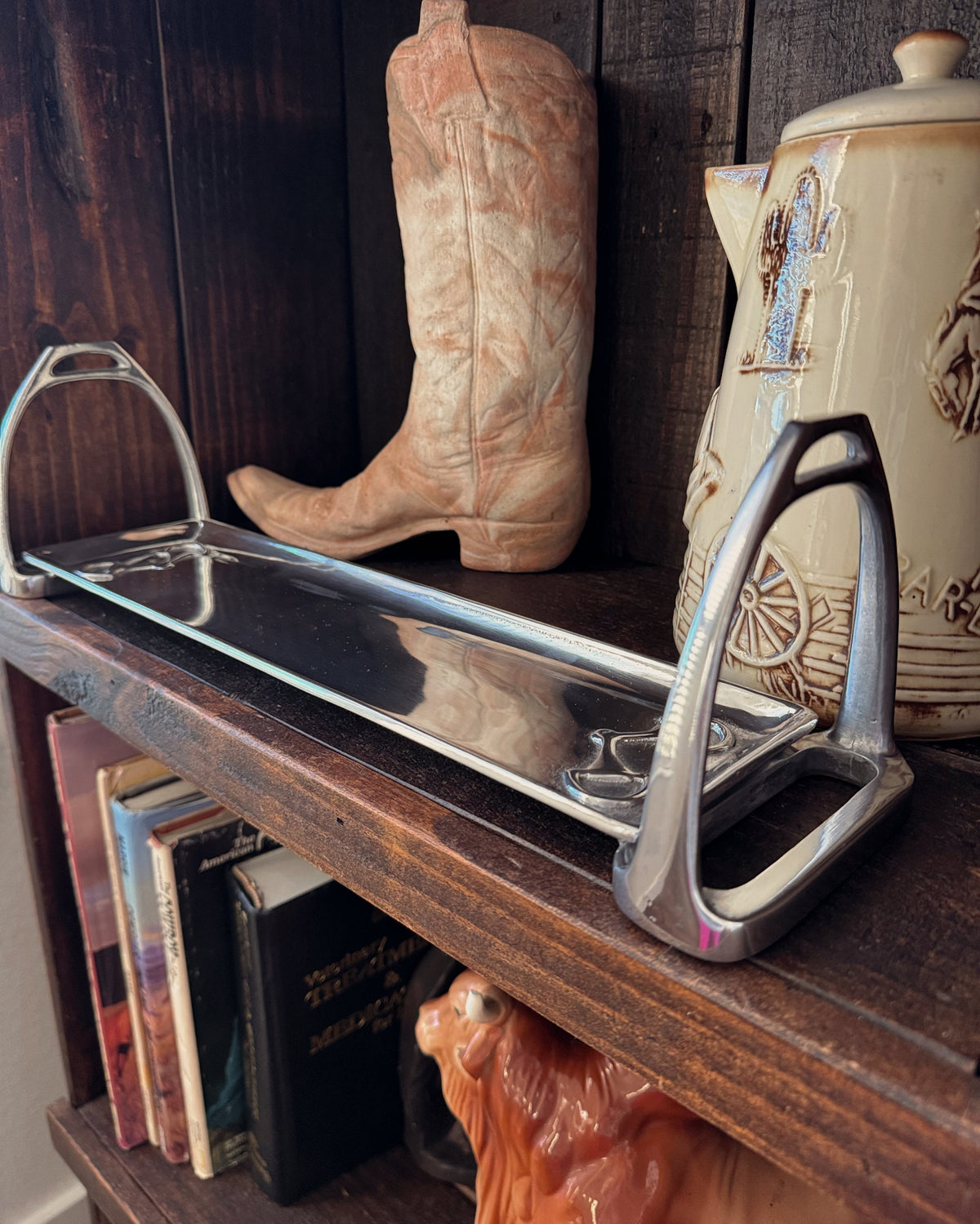 Old cowboy boot, metal stirrup, and ceramic pitcher on a wooden shelf.