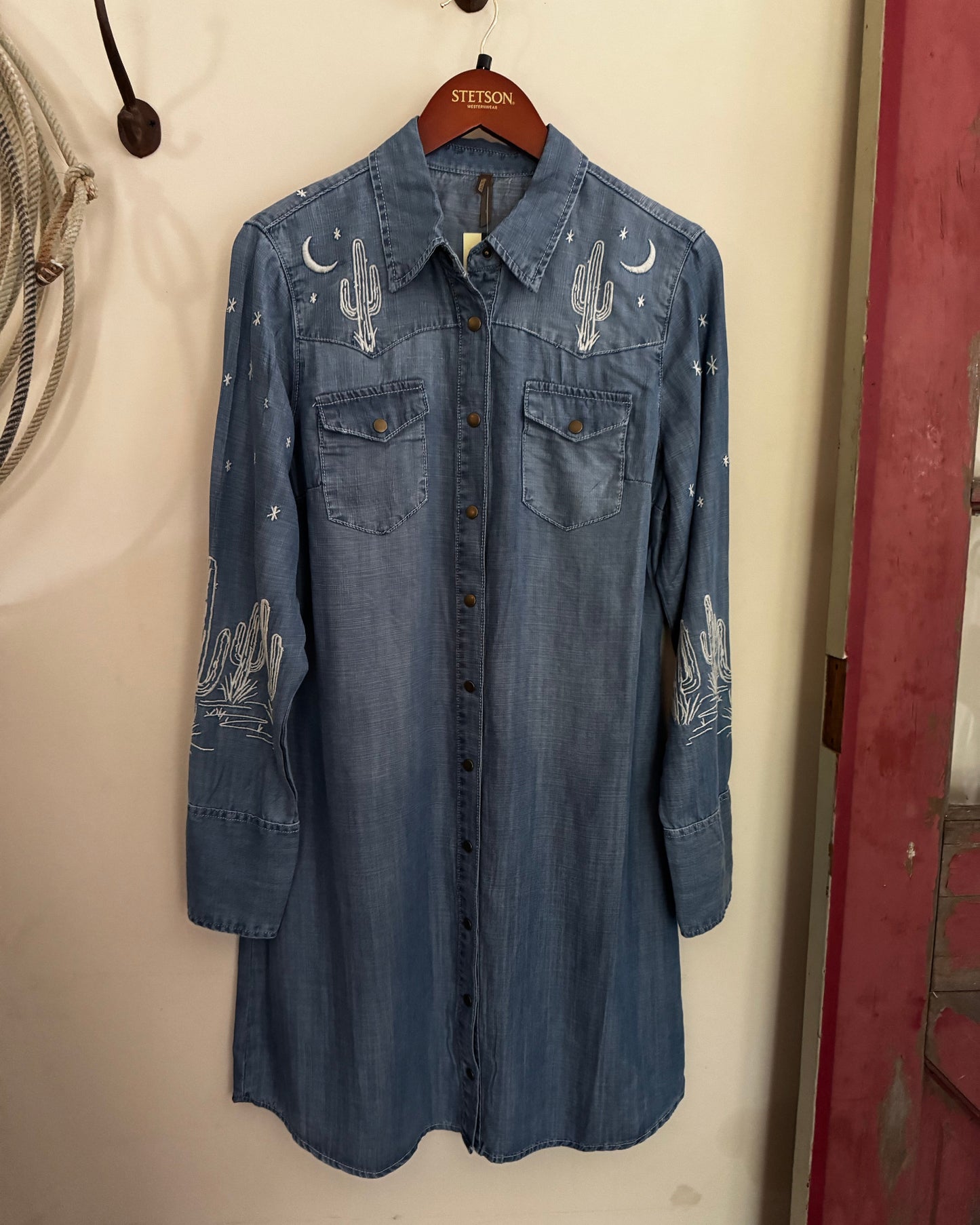 Blue denim shirt with cactus and moon designs on a hanger against a white wall.