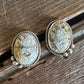 A pair of oval stud earrings with textured, crackled stone faces in cream and gold tones, set in a twisted silver-tone frame with small dangling beads, resting on a rustic wooden surface.