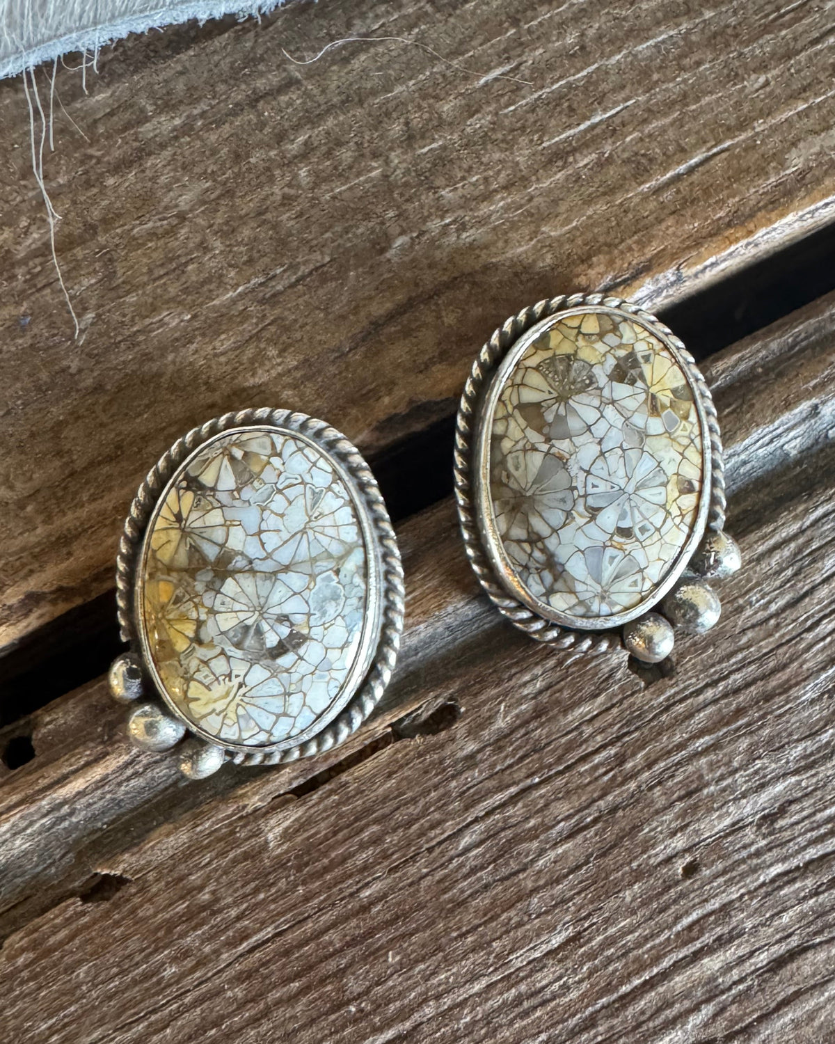 A pair of oval stud earrings with textured, crackled stone faces in cream and gold tones, set in a twisted silver-tone frame with small dangling beads, resting on a rustic wooden surface.