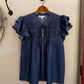 Blue blouse with ruffled sleeves on a wooden hanger against a beige wall.