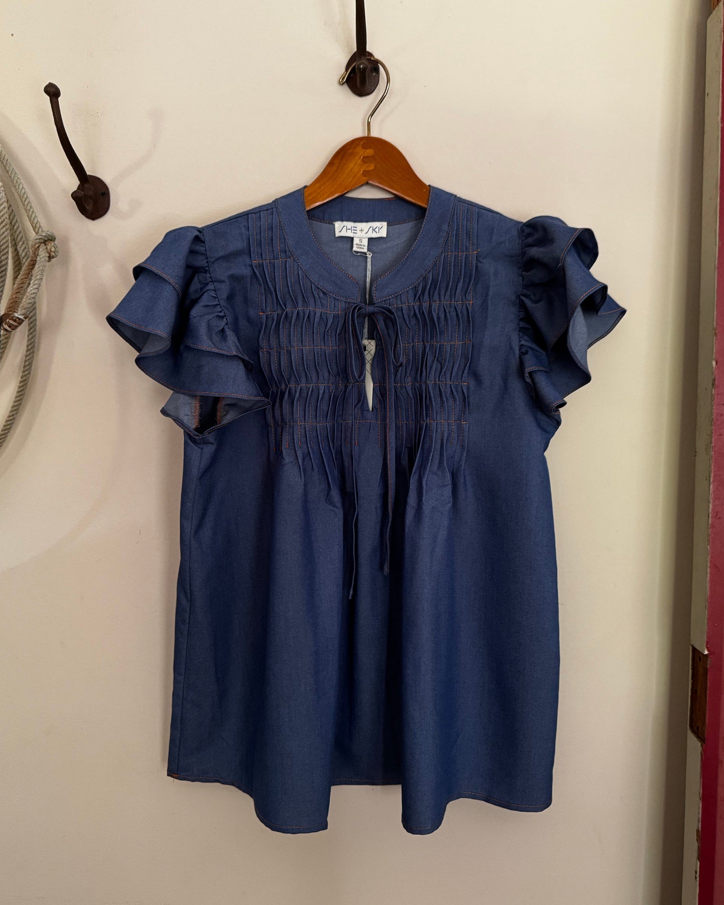Blue blouse with ruffled sleeves on a wooden hanger against a beige wall.