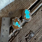 A pair of oval turquoise and brown stone earrings with silver-toned rope-style borders, displayed on a rustic wooden surface next to a metal ruler for scale.