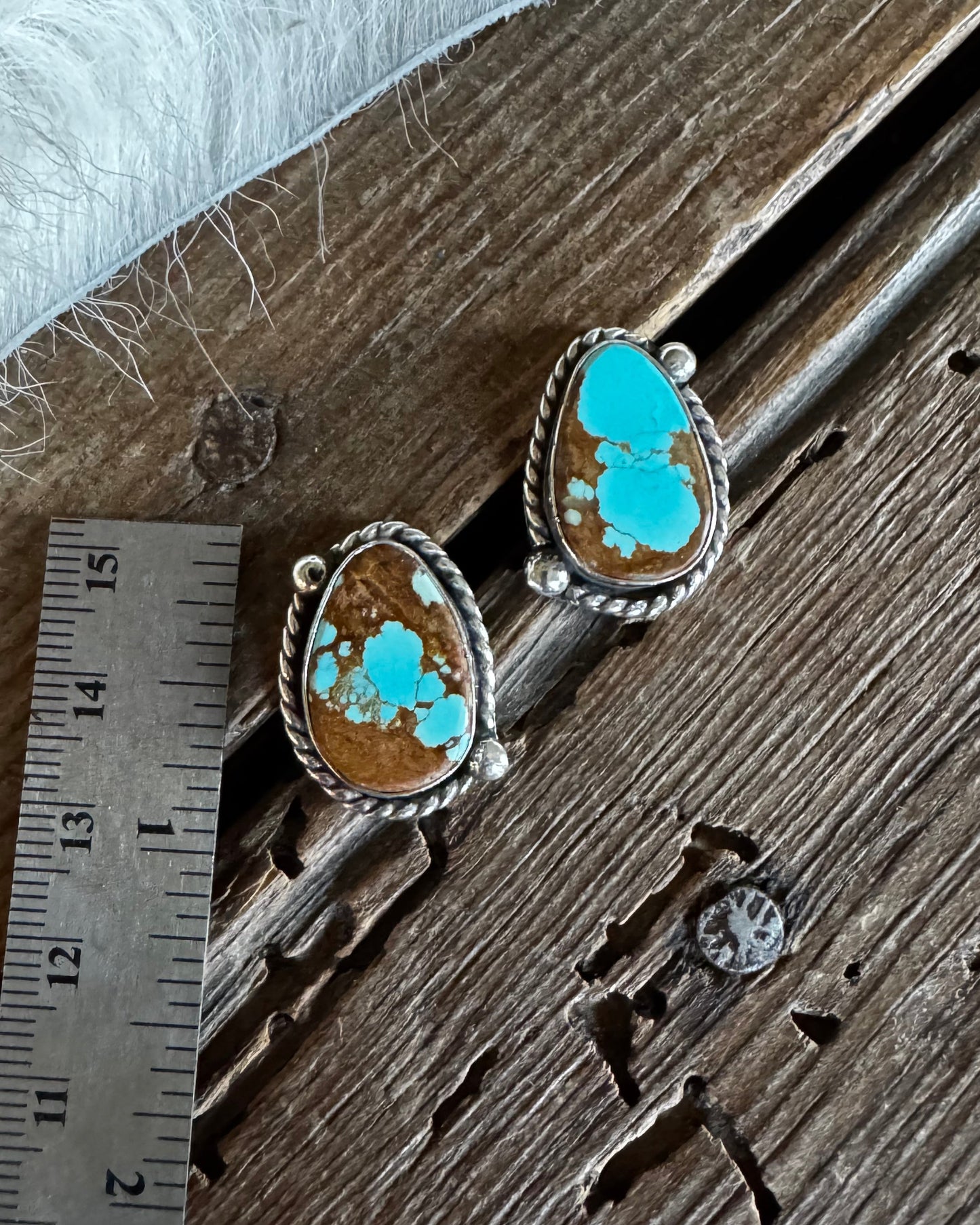 A pair of oval turquoise and brown stone earrings with silver-toned rope-style borders, displayed on a rustic wooden surface next to a metal ruler for scale.