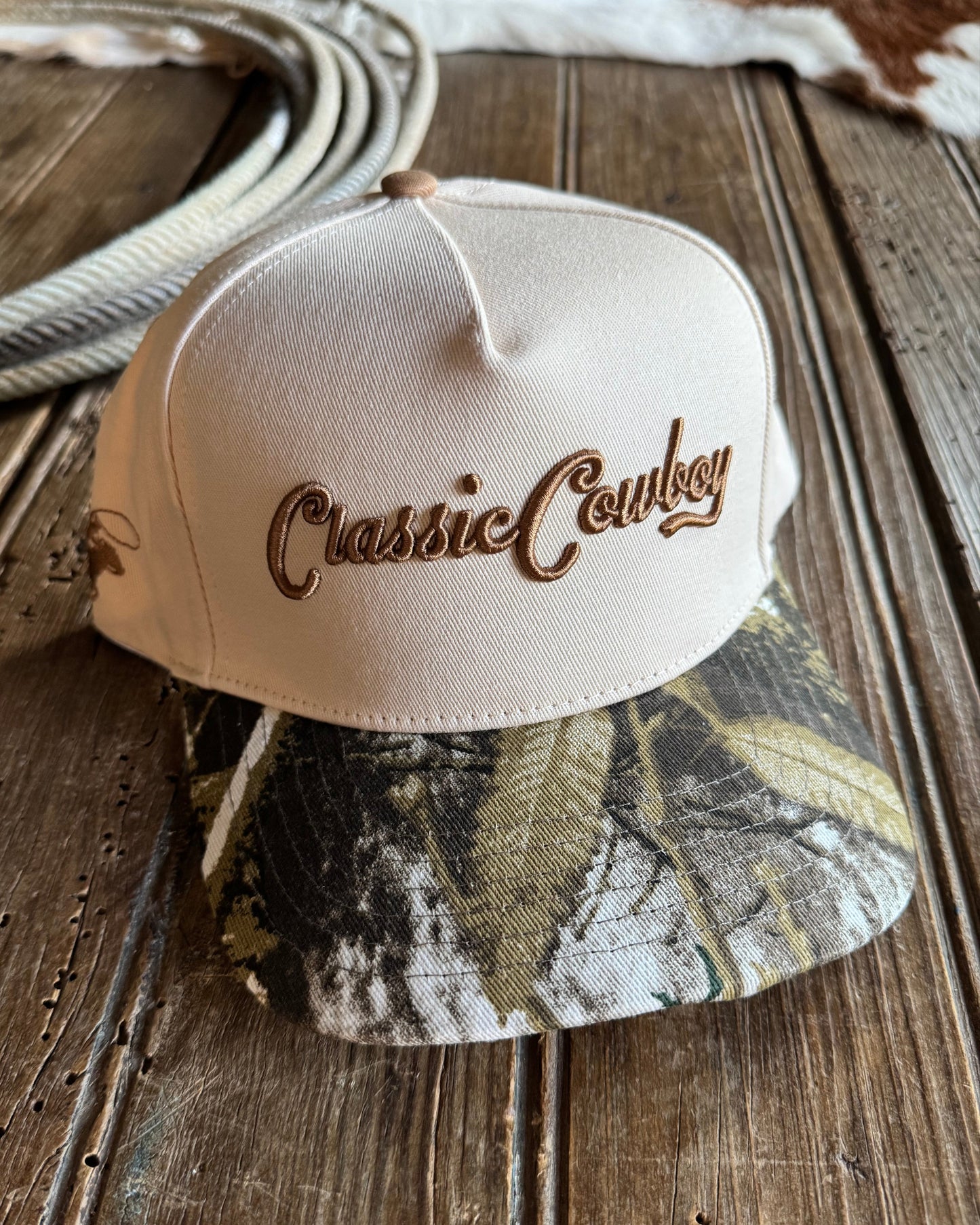 Beige and camouflage baseball caps with 'Classic Cowboy' branding on a wooden surface.