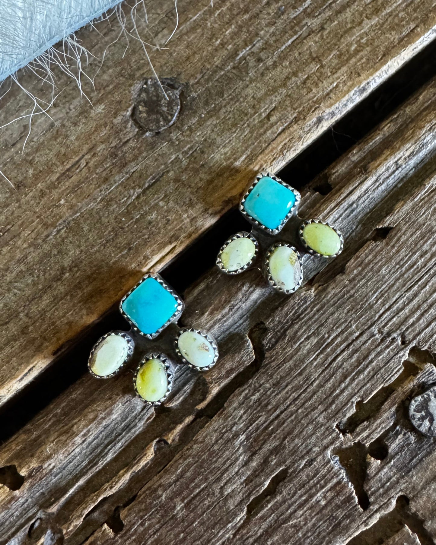 A pair of small silver-toned stud earrings with square turquoise-blue centers and oval milky-white stones, arranged on a rustic wooden surface with a white textured fabric at the top.