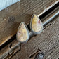 A pair of teardrop-shaped stud earrings with textured beige and white stone inlays set in silver, resting on a rustic dark wood surface.