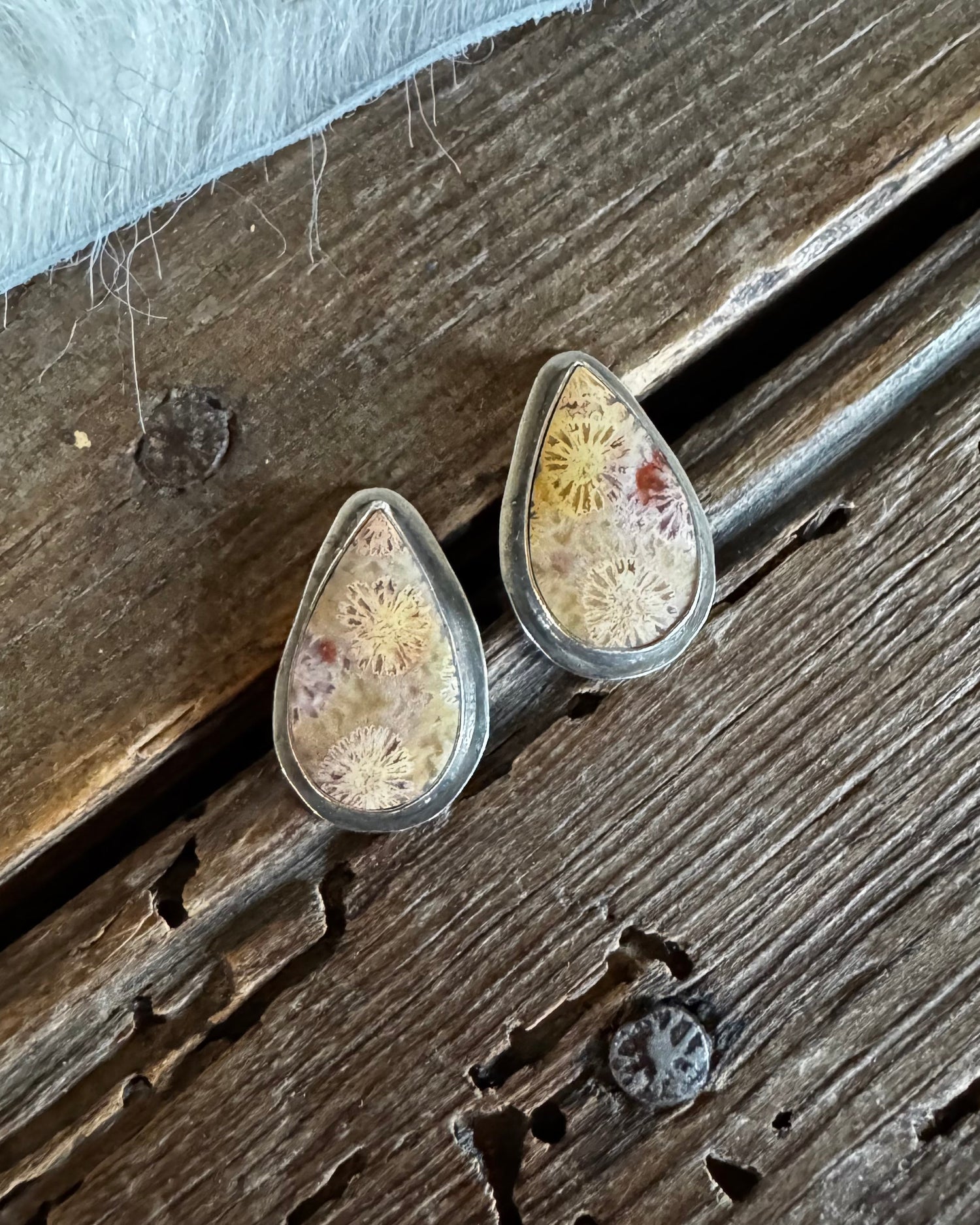 A pair of teardrop-shaped stud earrings with textured beige and white stone inlays set in silver, resting on a rustic dark wood surface.